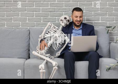 Young businessman with laptop and human skeleton sitting on sofa in office Stock Photo