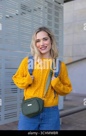 Woman standing holding backpack straps in yellow sweater on paved walkway by perforated panel Stock Photo