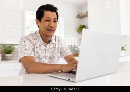 Asian man using laptop at home, smiling and working in bright kitchen Stock Photo
