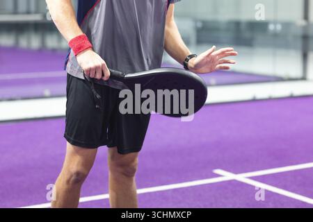 Male padel player in sportswear on purple court, gripping black racket and raising open hand Stock Photo