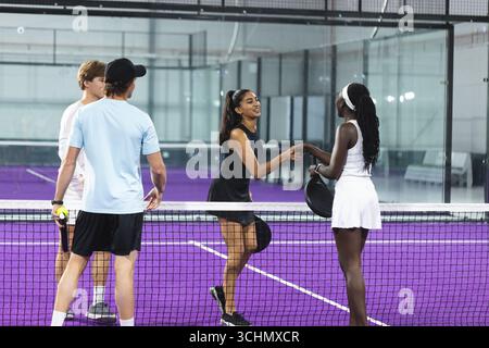 Diverse padel players in sportswear shaking hands at net on court in glass facility with rackets Stock Photo