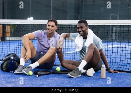 Sitting diverse male friends on blue court after playing pickleball with paddles, water bottles Stock Photo