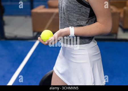 Female player holding yellow tennis ball on blue court wearing grey top white skirt and smartwatch Stock Photo