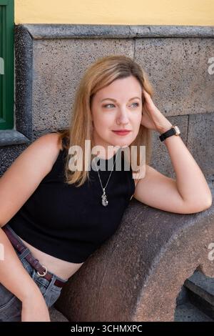 Young woman with blonde hair wearing black sleeveless top, grey jeans and a red belt while sitting outdoors against a textured stone bench Stock Photo
