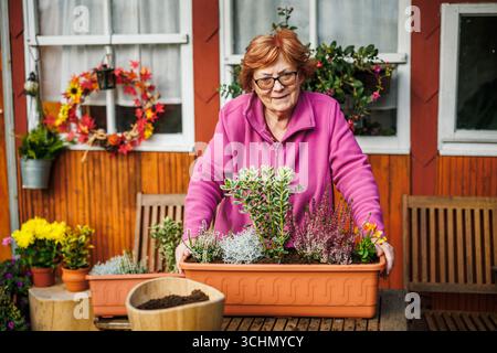 Autumn gardening. Happy senior woman arranging flowers and plant into flower pot for autumn garden and seasonal decoration. Stock Photo