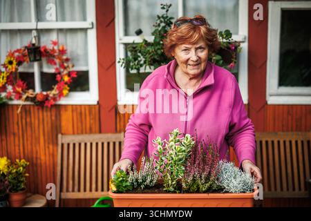 Happy senior woman arranging flowers and plant into flower pot for autumn garden and seasonal decoration. Fall gardening Stock Photo