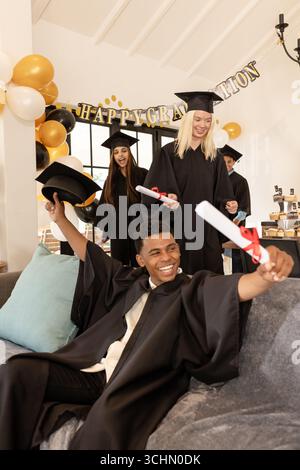 Diverse graduates celebrating with diplomas and caps, smiling joyfully at home party Stock Photo