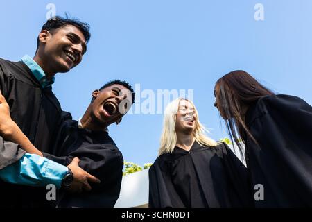 Diverse graduates in gowns celebrating achievements, laughing joyfully in garden, copy space Stock Photo