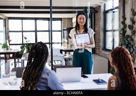 Asian woman presenting award to diverse female colleagues in modern office, smiling confidently Stock Photo