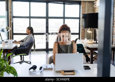 Focused diverse female professionals working on laptops in modern office, collaborating on projects Stock Photo