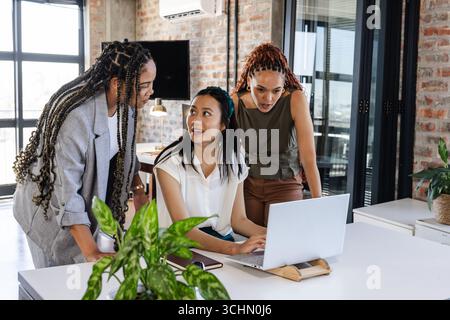 Collaborating in office, diverse female team sharing ideas around laptop enthusiastically Stock Photo
