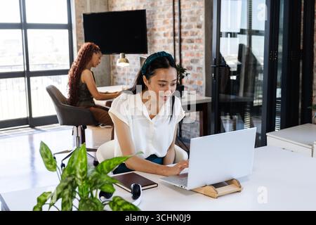 Asian woman working on laptop in modern office, smiling and focused Stock Photo