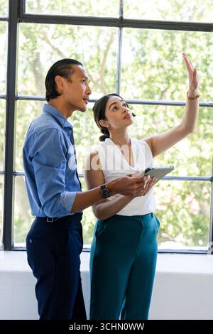 Discussing project details, business diverse professionals using tablet by large window, at office Stock Photo