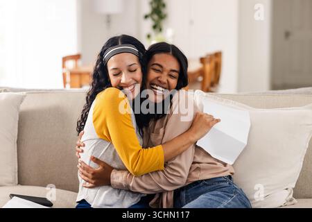 Two happy diverse female friends hugging on couch, celebrating good news at home Stock Photo