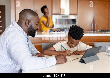 Father helping son with homework at kitchen table, mother talking on phone Stock Photo