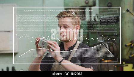 Examining metalworker in apron and goggles holding metal component in workshop, with data overlay Stock Photo