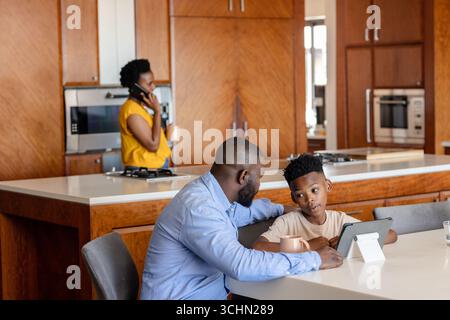 Father and son using tablet at kitchen table while mother talks on phone Stock Photo