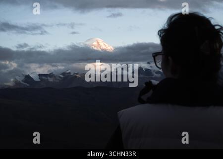 The silhouette of a person looking out at Mount Everest, its snowy peak glowing in sunset light beneath a lenticular cloud. Stock Photo