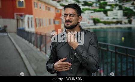 Young man in glasses and gray shirt stands on promenade near seaside holding throat with distressed expression against blurred beach backdrop Stock Photo