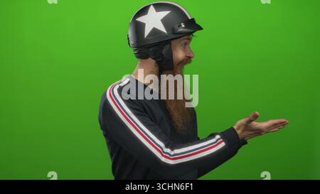 Young man with long beard wearing black motorcycle helmet and striped sleeve sweater presents open palms in green studio; joy. Stock Photo