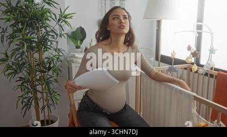 Pregnant woman sitting in a light-filled nursery cooling herself with a paper fan beside a crib surrounded by baby decor and plants, looking thoughtfu Stock Photo