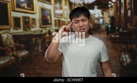 Young chinese man making a puzzled gesture indoors at a museum with ornate art in the background, wearing a white shirt, creating a humorous or confus Stock Photo