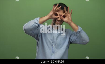 Smiling young hispanic man in casual blue shirt holding hands like binoculars over eyes against isolated green background. Stock Photo