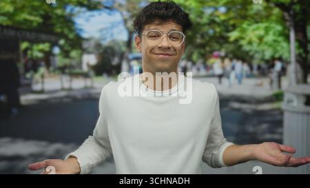 Man wearing round glasses and a white sweater shrugs both shoulders amid passersby on a sunny urban street; uncertainty. Stock Photo