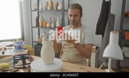 Man with long blond hair and apron holds red smartphone while seated at pottery wheel and examines a clay vase in studio; creative focus. Stock Photo