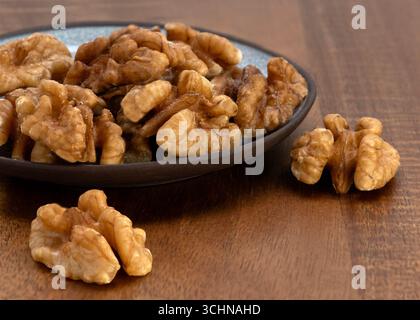 Close-up of de-shelled walnuts on a ceramic plate with a few scattered on a wooden table, perfect for food, nutrition, and healthy lifestyle concepts. Stock Photo