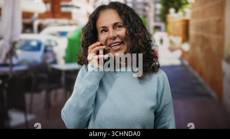 Middle-aged woman at outdoor cafe talking happily on phone with warm expression in a lively terrace setting. Stock Photo