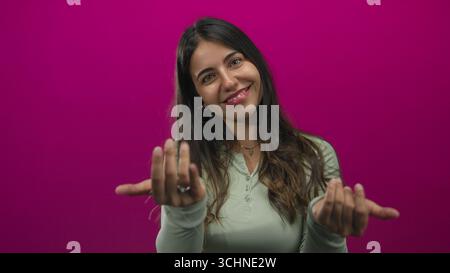 Hispanic woman beckons with open hands in a pink studio with a bright friendly smile; warm invitation. Stock Photo