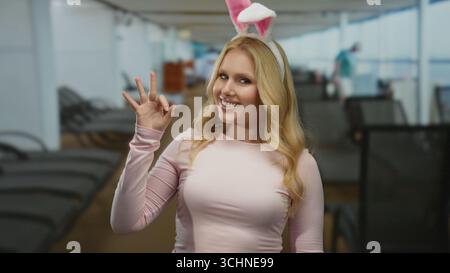 Young blonde woman with bunny ears makes an ok gesture while smiling on a cruise ship deck, exuding a funny and playful vibe amidst a relaxing environ Stock Photo