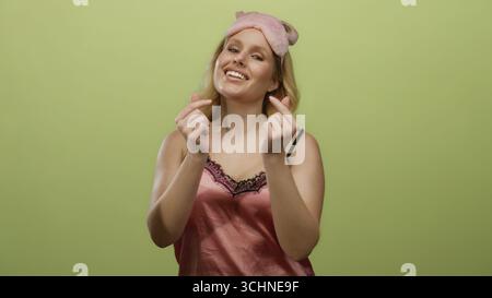 Young blonde woman in pink nightwear makes heart gesture with hands against yellow background wearing a playful sleeping mask, conveying a sense of af Stock Photo