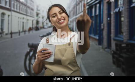 Woman waitress in beige apron holds notepad and pen and shows middle finger while smiling on city street; defiance. Stock Photo