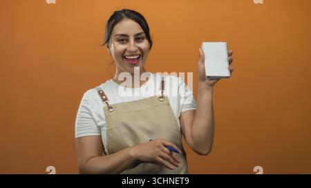 Woman in apron holding a notepad and pen, smiling and showing her hand in an orange studio; friendly service. Stock Photo