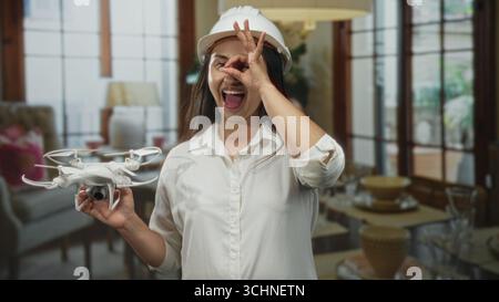 Woman holding a white drone and making ok sign to eye in building with hardhat on and smiling; joyful innovation. Stock Photo