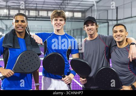 Diverse male friends in sportswear and standing with padel net on padel court, holding rackets Stock Photo