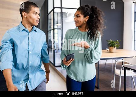 Diverse colleagues discussing project while walking in modern office, holding smartphone Stock Photo
