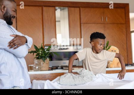 Father watching son folding laundry at home, bonding in kitchen area Stock Photo