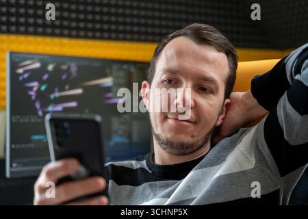 Man relaxing and smiling while looking at smartphone in modern tech workspace, suggesting casual moment during work break Stock Photo