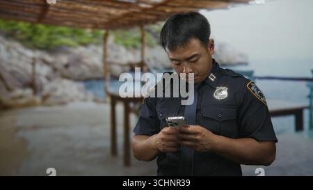 Chinese policeman holds smartphone in both hands beside building walkway under wooden canopy wearing uniform and radio gear; determination. Stock Photo