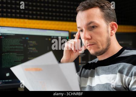 Focused man reading printed documents in software development workspace with code on monitors, reflecting analytical work in tech environment Stock Photo