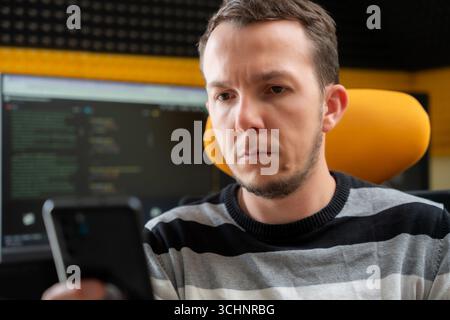 Man looking at smartphone with serious expression in programming workspace, reflecting concentration or tech-related troubleshooting Stock Photo