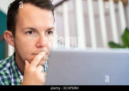 Thoughtful Man Holding Chin Looking At Laptop Screen At Home Office Remote Work Stock Photo