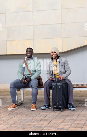 Diverse male friends sitting on bench at courtyard using smartphone with headphones Stock Photo