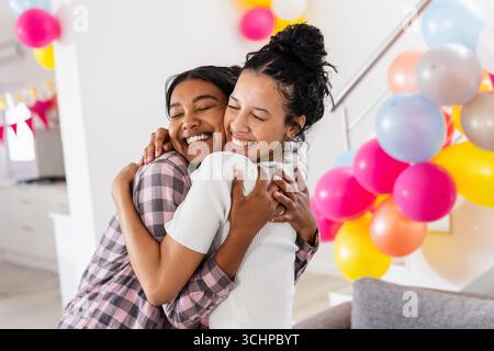 Two diverse female friends hugging joyfully at home surrounded by colorful balloons Stock Photo