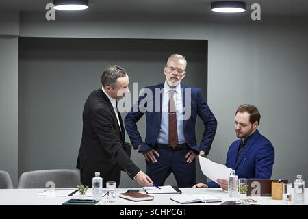 Three Caucasian middle aged businessmen discussing documents in modern office meeting room, one man standing with hands on hips while two others reviewing papers at conference table Stock Photo