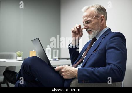 Senior Caucasian man sitting at desk using laptop, adjusting eyeglasses while focusing on screen, business professional working in modern office environment Stock Photo