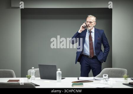 Mature Caucasian man in suit standing in modern office talking on smartphone, wearing suit and tie, looking away from laptop on conference table, holding hand in pocket Stock Photo